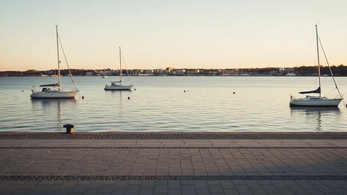 Ruhige Bucht der Ostsee zur goldenen Stunde mit einigen festgemachten Segelbooten, breite Steinpromenade im Vordergrund, niedrige Stadtpanorama im Hintergrund verschwommen