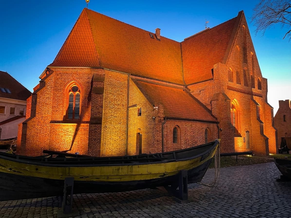 Oświetlony nocą ceglany budynek Muzeum Rybołówstwa w Helu z drewnianą łodzią na pierwszym planie.