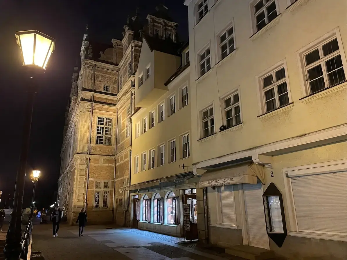 نزهة مسائية على ضفاف Długie Pobrzeże في غدانسك تظهر المنازل التاريخية والواجهة المضاءة لبوابة Zielona Brama على طراز عصر النهضة في البلدة القديمة.