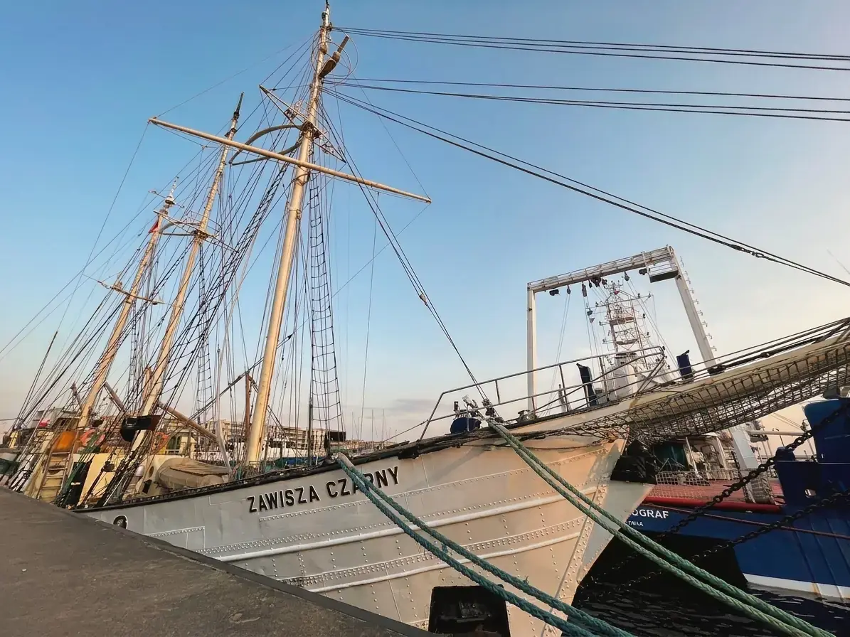 Weißes Segelschiff Zawisza Czarny am Betonkai im Hafen von Gdynia mit hohen Masten und dicken Festmacherleinen im Vordergrund.